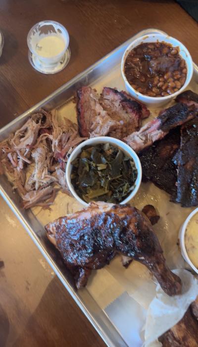 Barbecue platter with pulled pork, ribs, smoked chicken, collard greens, baked beans, and mac and cheese served on a metal tray with barbecue sauce on the side.