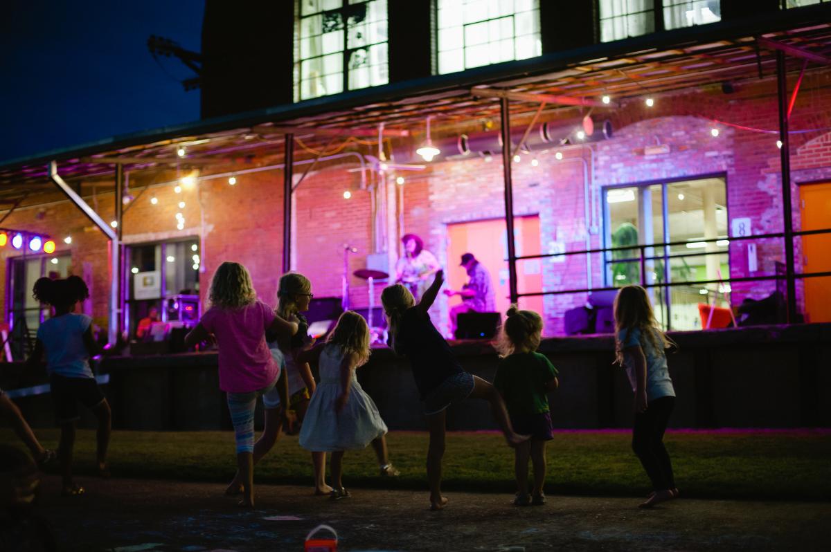 Children dance joyfully in front of a vibrant outdoor stage in Huntsville with live musicians. Strings of lights and a warm brick backdrop create a festive, lively atmosphere.