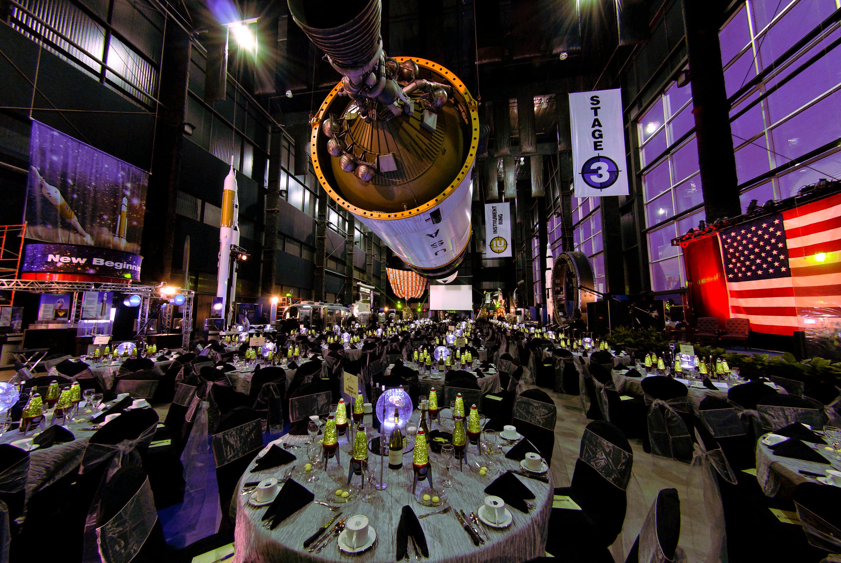 Rocket Center meeting space with elegant tables under Saturn V rocket and American flag backdrop with colorful lighting