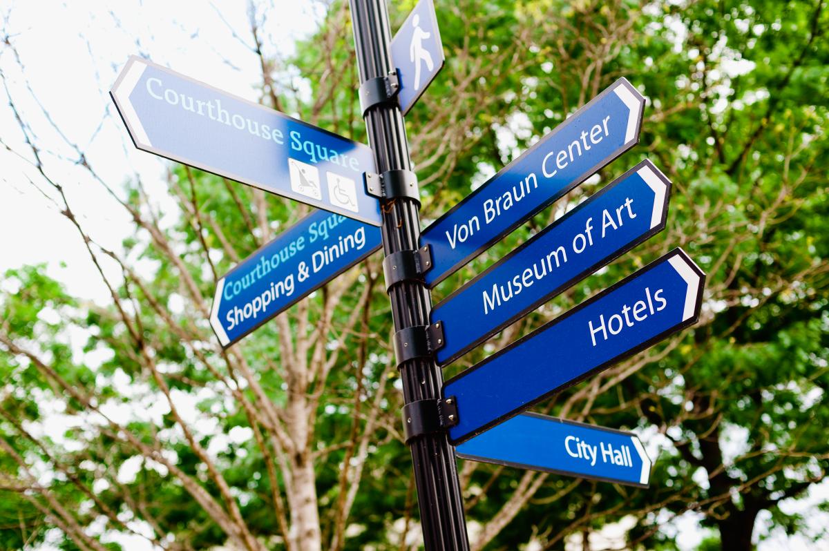 Blue directional signs in downtown Huntsville point to Courthouse Square, the Von Braun Center, the Huntsville Museum of Art, hotels, and City Hall, with trees visible in the background.