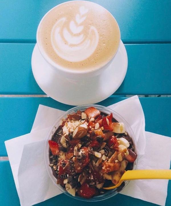 Latte with leaf foam art beside an acai bowl topped with fruit and nuts on a blue table in Huntsville, creating a fresh and inviting scene.