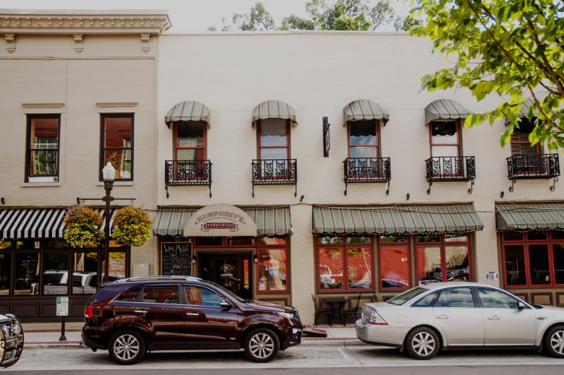 Two-story beige building in Huntsville with green-striped awnings and wrought iron balconies, with cars parked out front and vibrant trees creating a quaint feel.