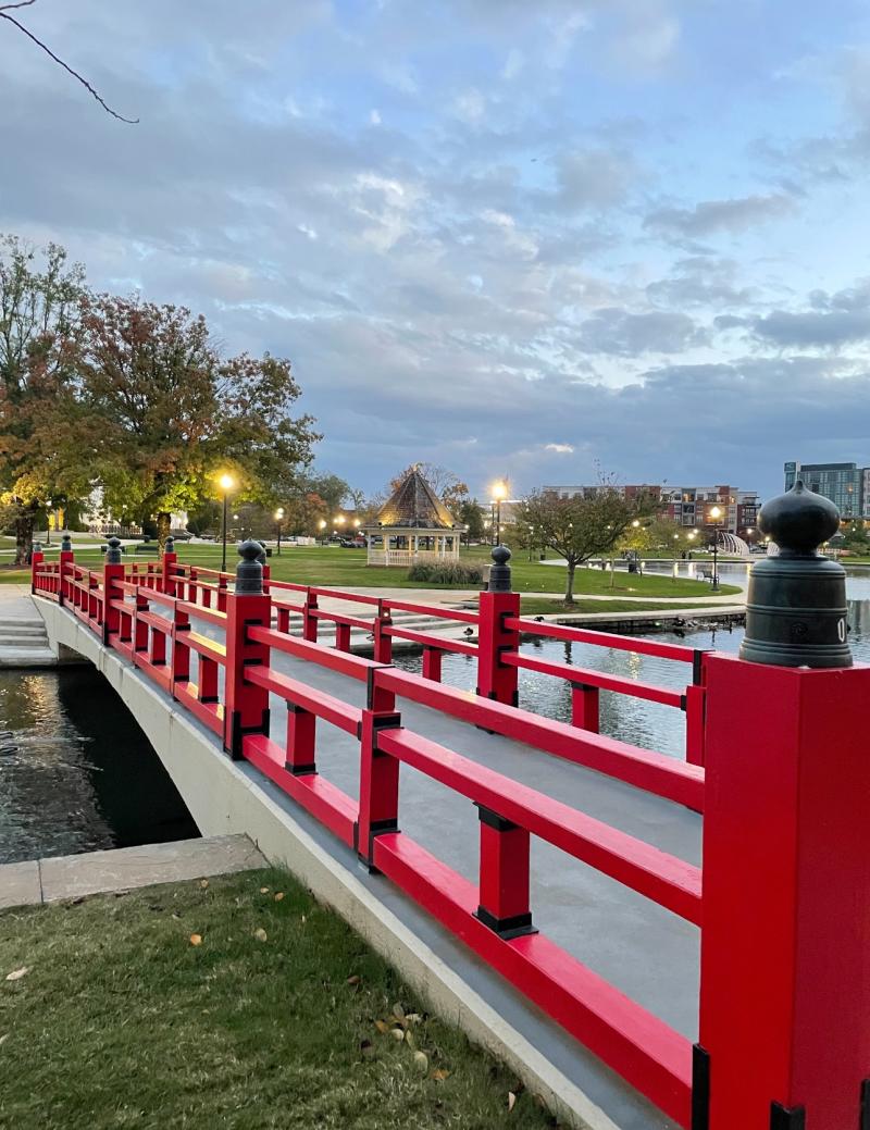 Red pedestrian bridge in Huntsville’s Big Spring International Park at sunset with water, gazebo, and downtown lights