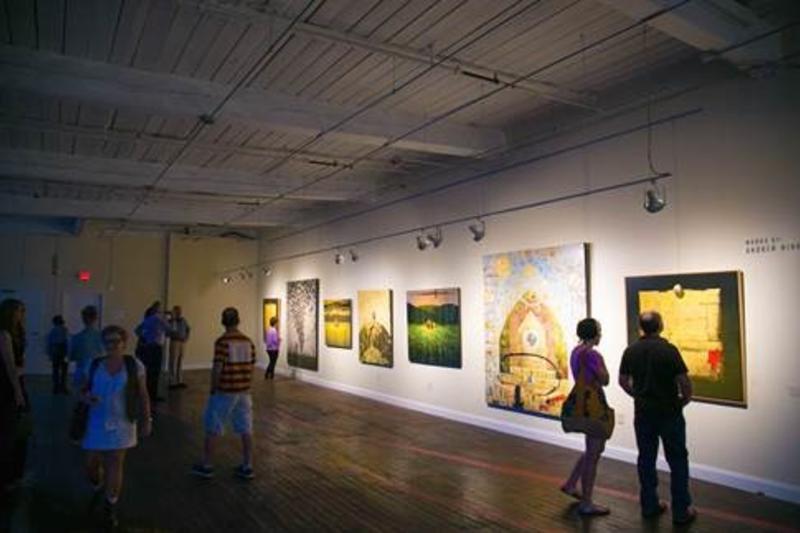 Dimly lit Huntsville art gallery with people viewing colorful paintings on a white wall. The wood floor and high ceiling create an open, reflective atmosphere.
