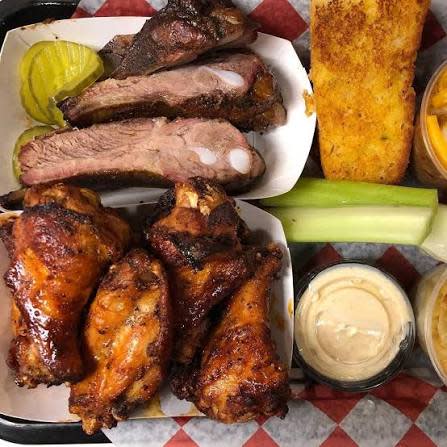 Barbecue platter from Moe’s BBQ featuring smoked ribs, chicken wings, cornbread, pickles, celery, and dipping sauces served in baskets.
