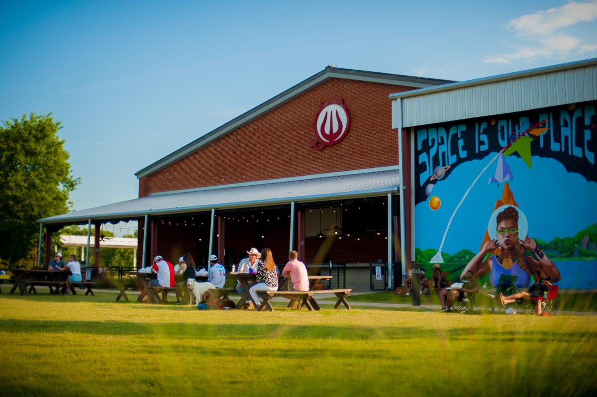 People sit on picnic benches outside a red brick brewery in Huntsville, featuring a large mural reading “Space Is Our Place” in a relaxed, sunny setting.