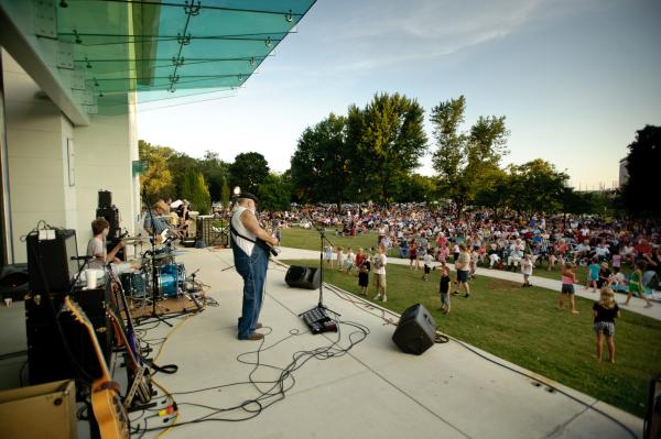 Live band performing on an outdoor stage as a large crowd gathers in a Huntsville park during a summer concert.