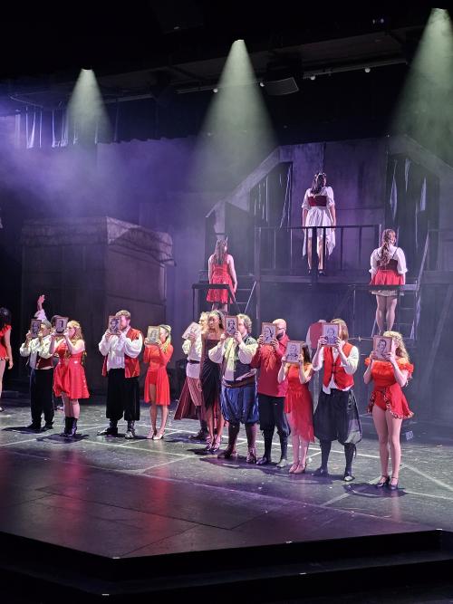 Cast members perform on stage during a Theatre Huntsville production, dressed in red, white, and black costumes while holding illustrated portraits. The ensemble stands beneath dramatic stage lighting in front of a multi-level set as part of a musical performance.