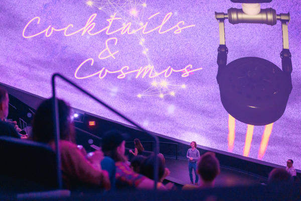 Guests gather inside the INTUITIVE® Planetarium at the U.S. Space & Rocket Center during the Cocktails & Cosmos event. A presenter stands beneath a colorful dome projection featuring stars, constellations, and event branding while attendees watch from theater-style seating with drinks in hand.