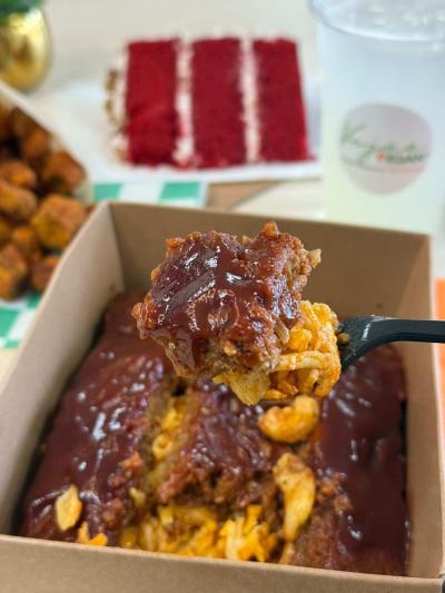 Close-up of a vegan barbecue mac and cheese dish from Vujee Vegan topped with barbecue sauce, with a slice of red velvet cake and a drink blurred in the background.